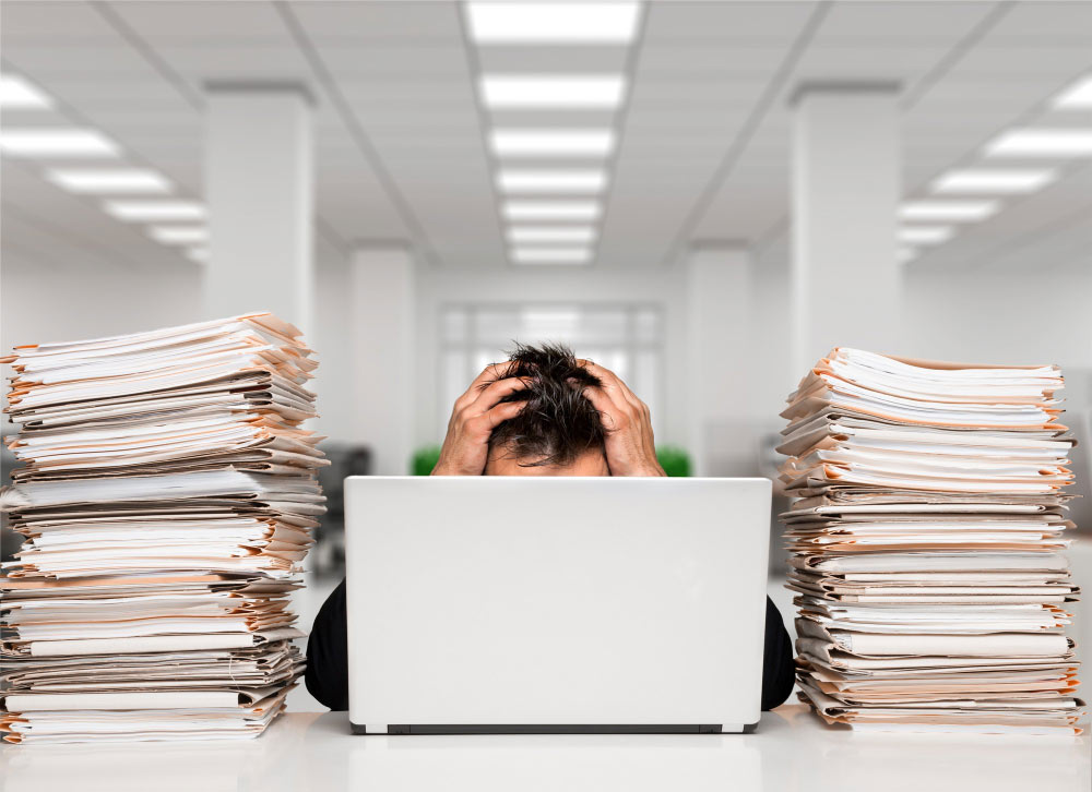 Persona frente a un computador con montones de documentos a los lados, reflejando la necesidad de optimizar procesos.